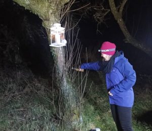 Nightime in orchard. Michelle dressed in blue waterproof jacket and red hat pouring cider on an apple tree lit by a candle lantern