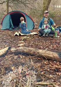 A father and his two young sons sitting on the ground having a bedtime drink sitting beside a campfire in front of their small tent in the woods