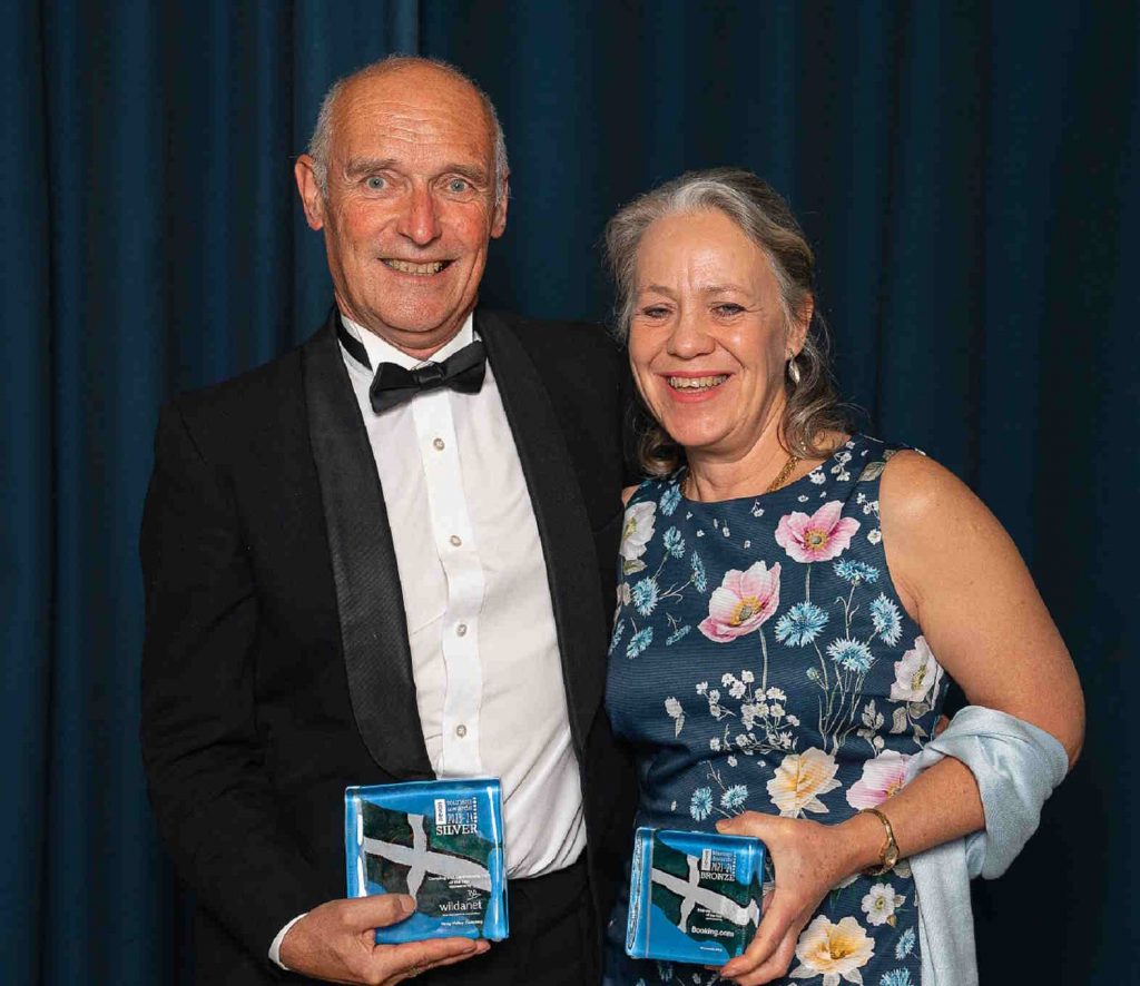 Michelle & Kevin holding their Devon Tourism Awards 2023 for the B&B and Campsite.  Kevin is wearing a DJ and Michelle a blue flowery shift dress