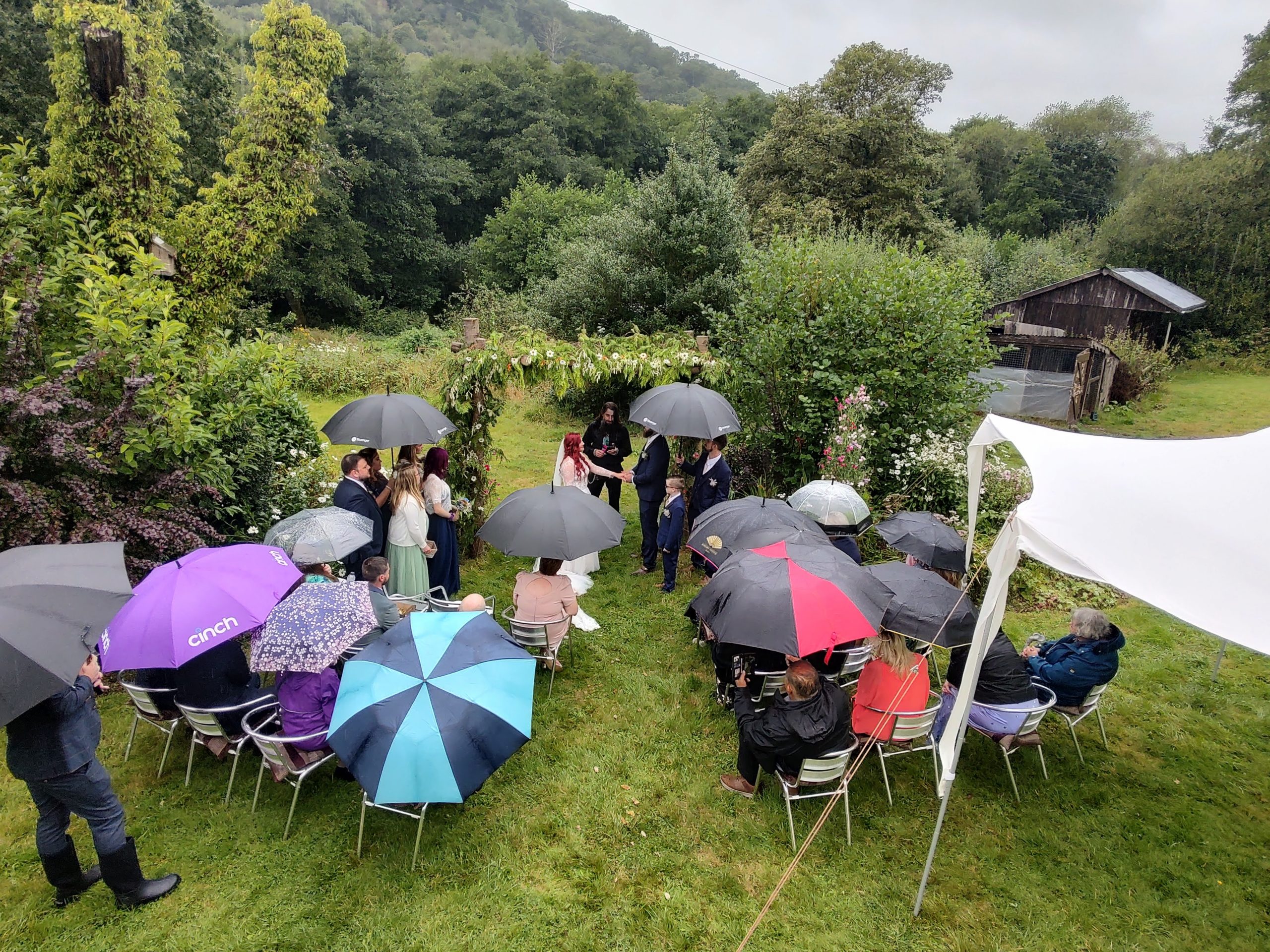 A wedding blessing in the garden at Wray Valley.  It is raining and teh guests are seated sheletering under large umbrellas.  The bride is in a white long dress and has long red hair.  The union is being blessed by a male celebrant. The couple and the celebrant are standing beneath a floral pergola 