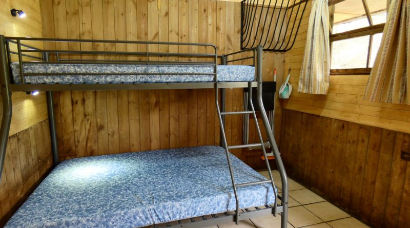 Inside of one of the Stables Eco-Bothies. Room is clad in pine and has a cream tiled floor. Shows triple bunk) double and single) with mattresses. Window with curtains to right. Original hay rack on wall