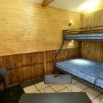 Inside of one of the Stables Eco-Bothies Accommodation Units. Room is clad in pine and has a cream tiled floor. Shows triple bunk) double and single) with mattresses, black ikea fold away table and 3 chairs. Small Outbacker black wood burning stove in corner