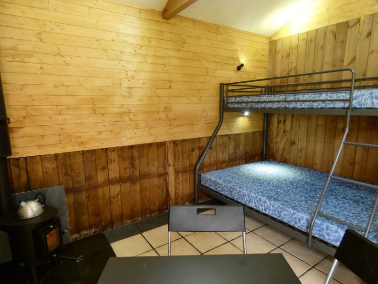 Inside of one of the Stables Eco-Bothies Accommodation Units. Room is clad in pine and has a cream tiled floor. Shows triple bunk) double and single) with mattresses, black ikea fold away table and 3 chairs. Small Outbacker black wood burning stove in corner