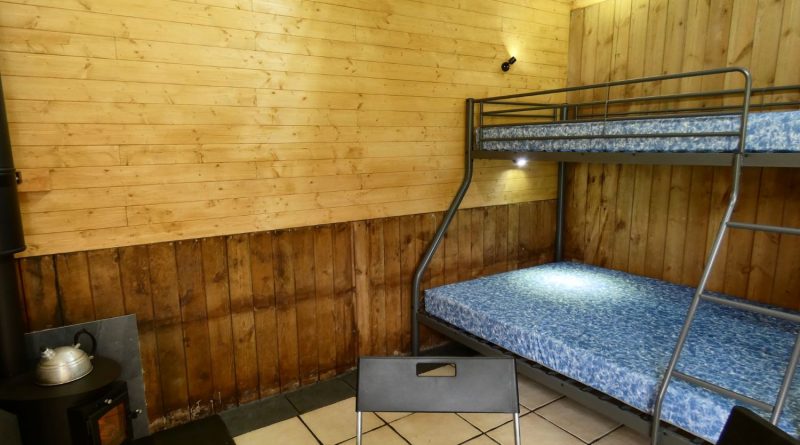 Inside of one of the Stables Eco-Bothies Accommodation Units. Room is clad in pine and has a cream tiled floor. Shows triple bunk) double and single) with mattresses, black ikea fold away table and 3 chairs. Small Outbacker black wood burning stove in corner