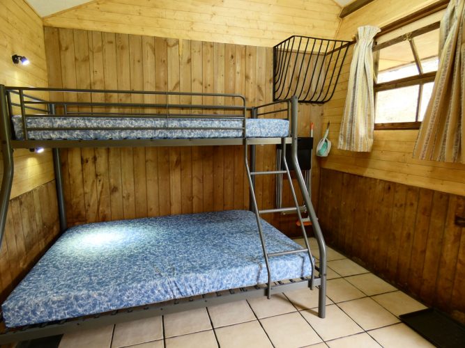 Inside of one of the Stables Eco-Bothies. Room is clad in pine and has a cream tiled floor. Shows triple bunk) double and single) with mattresses. Window with curtains to right. Original hay rack on wall