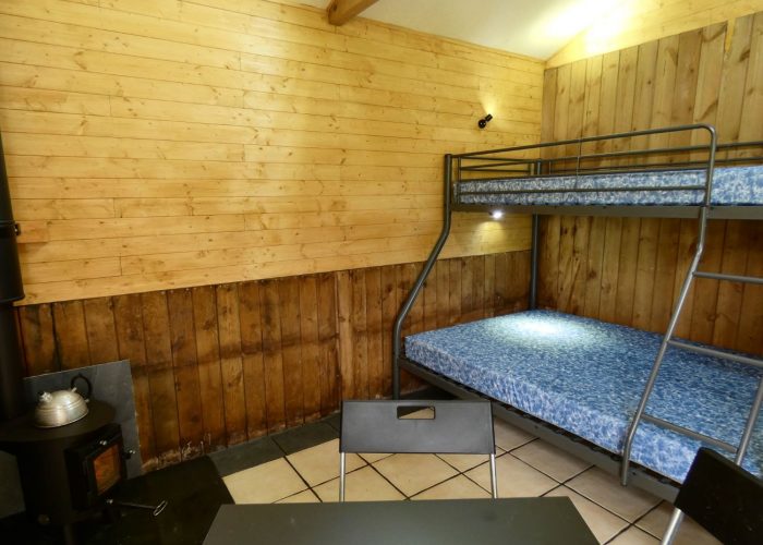 Inside of one of the Stables Eco-Bothies Accommodation Units. Room is clad in pine and has a cream tiled floor. Shows triple bunk) double and single) with mattresses, black ikea fold away table and 3 chairs. Small Outbacker black wood burning stove in corner